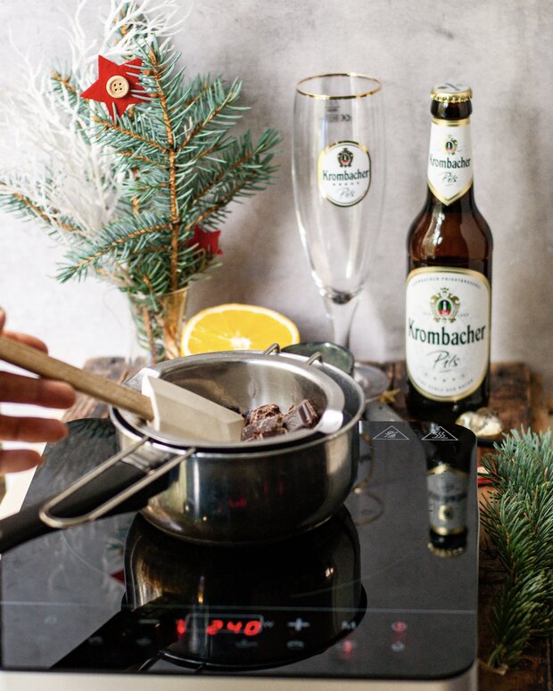 Ein Kochtopf mit geschmolzenem Schokoladeninhalt auf einem Induktionsherd. Daneben stehen ein Krombacher Pils Bier und ein Glas mit dem Krombacher Logo.