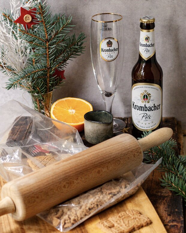 Ein Krombacher Pils Bier in einer Flasche und ein Krombacher Glas stehen neben einer Orange, einem Teelicht und einem Nadelbaum. Im Vordergrund liegt ein Nudelholz auf einem Holzbrett, sowie zerbröseltem Lebkuchen.