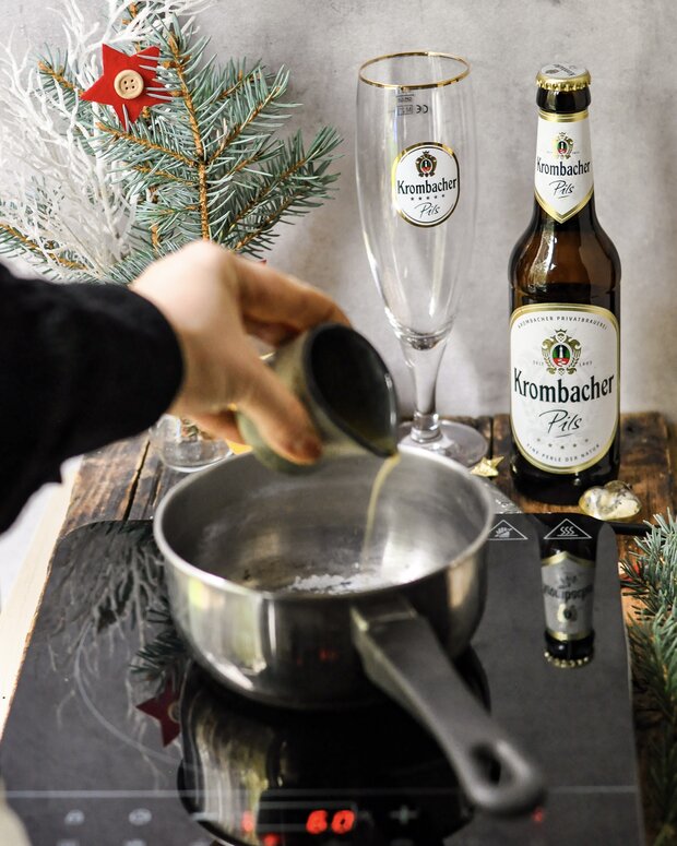 Eine Hand gießt eine Flüssigkeit aus einer kleinen Dose in einen silbernen Kochtopf auf einem Induktionsherd. Im Hintergrund stehen ein Krombacher Pils Bier und ein dazugehöriges Glas.
