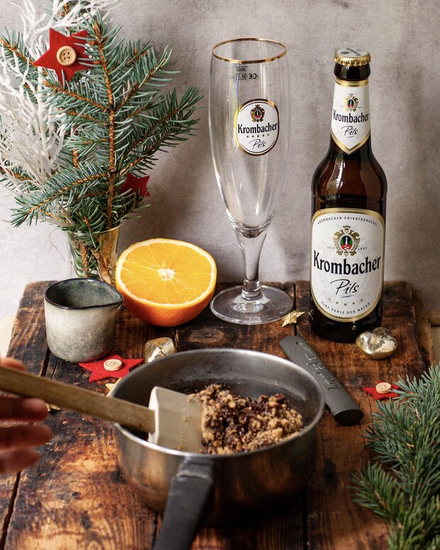 Ein Topf mit Teig und einem Spatel steht auf einem Holztisch. Hinter dem Topf sind eine Flasche und ein Glas Krombacher Pils sowie eine Orange und Weihnachtsdekoration.