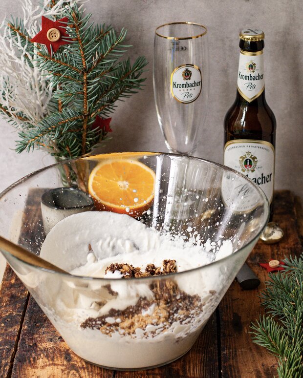 Eine große Glasschüssel mit einer Mischung aus Sahne und den Lebkuchen steht auf einem Holztisch. Im Hintergrund sind ein Krombacher Bier, ein Glas, eine Orange und ein Tannenzweig zu sehen.
