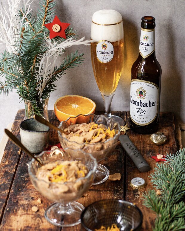 Zwei Schalen mit Lebkuchen-Mousse, garniert mit Orangenschalen. Im Hintergrund steht eine Flasche 'Krombacher' und Weihnachtsdekoration.