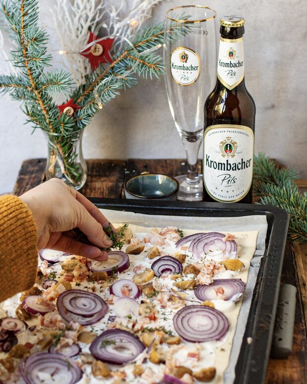 Eine Person mit einer Hand bestreut einen belegten Flammkuchen mit Zwiebeln und Kräutern. 