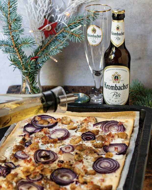 Ein Blech Flammkuchen mit roten Zwiebeln und Kräutern wird mit Olivenöl beträufelt.