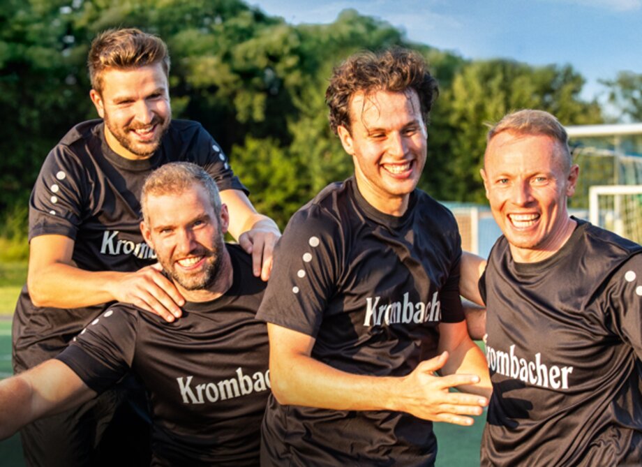 Vier Männer in schwarzen Krombacher-Trikots lachen und umarmen sich auf einem Fußballfeld im Freien.