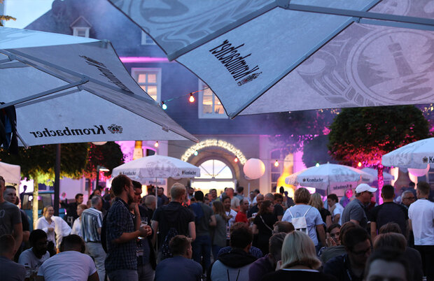 Festliche Abend-Szene mit vielen Menschen vor der Krombacher Brauerei. Stimmungsvolles Licht färbt die Umgebung ein.
