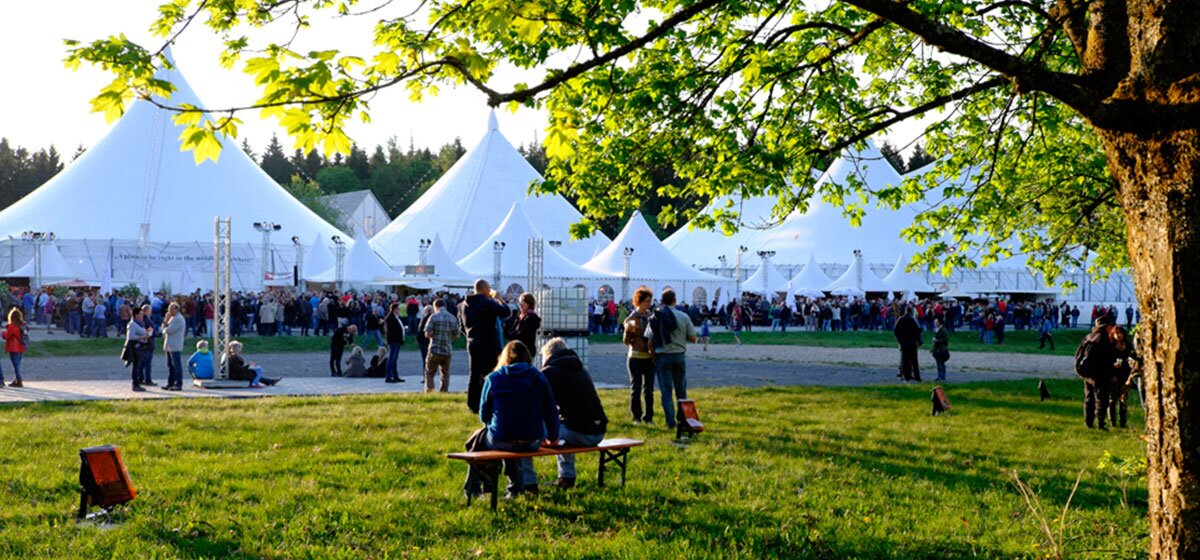 Viele weiße Festzelte mit spitzen Dächern, auf einer grünen Wiese, mit vielen Menschen davor.