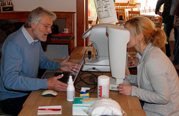 Ein Arzt und eine Patientin sitzen an einem Holztisch. Die Patientin schaut durch ein medizinisches Gerät zur Augenuntersuchung.