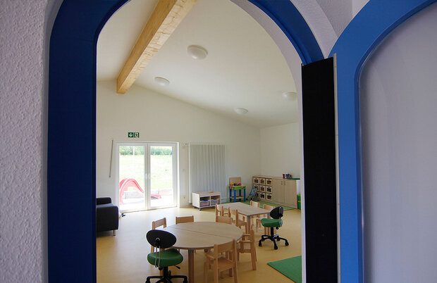 Abbildung eines Raumes der Krombärchen Kita. Es ist ein Holz-Tisch mit Stühlen zu sehen, sowie Regalen mit Spielzeug.