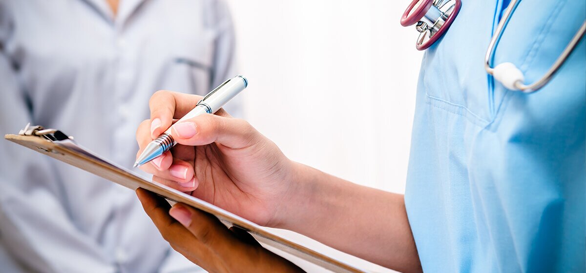 Hand einer weiblichen, medizinischen Fachkraft in blauer Kleidung mit Stethoskop, die auf einem Klemmbrett Notizen macht.