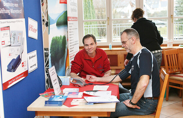 Zwei Männer sitzen an einem Tisch mit Infomaterial. Einer der Männer untersucht mit einem Blutdruckmessgerät den anderen Mann.
