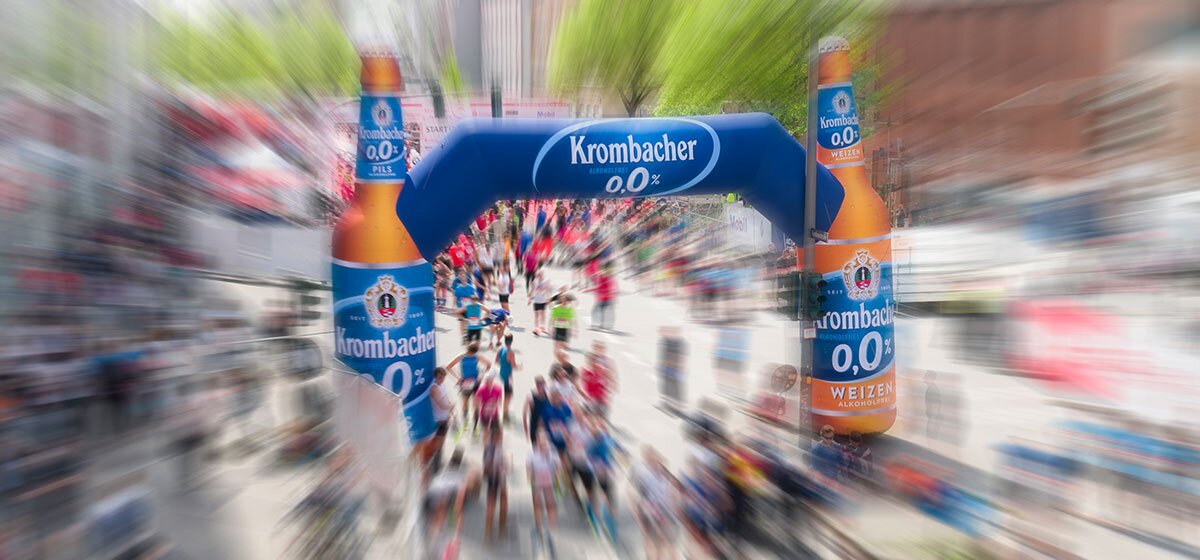 Läufer passieren einen blauen Krombacher o,0% Werbebogen bei einem Marathon-Lauf.