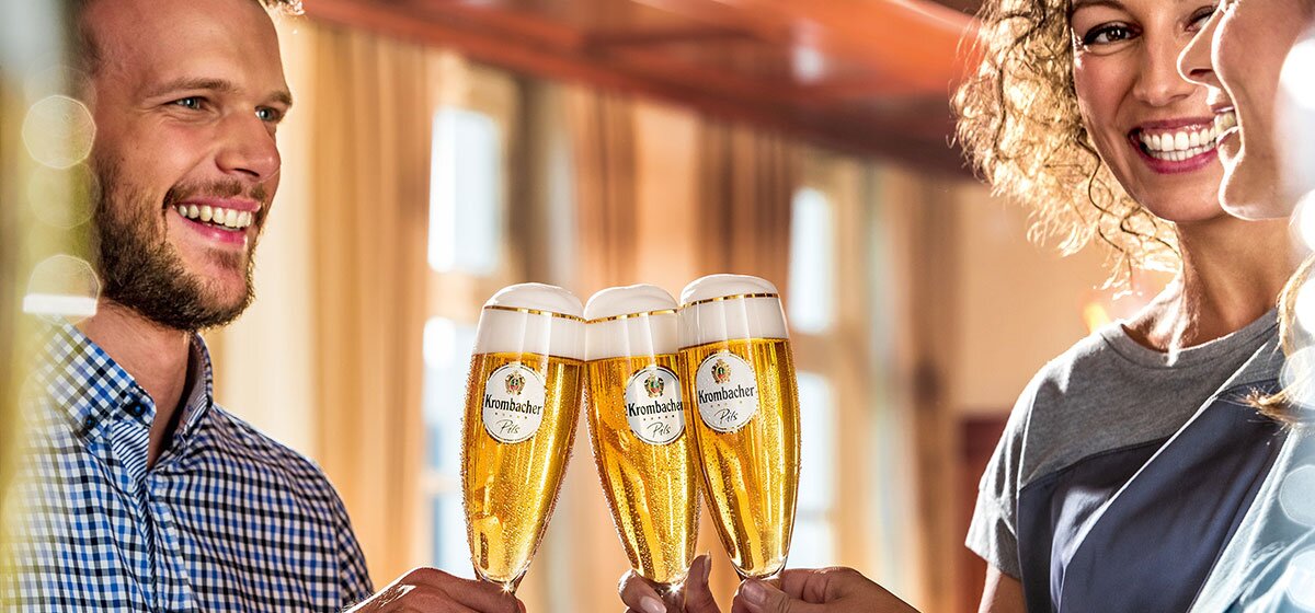 Zwei Frauen und ein Mann stoßen mit einem Krombacher Pils Glas an.