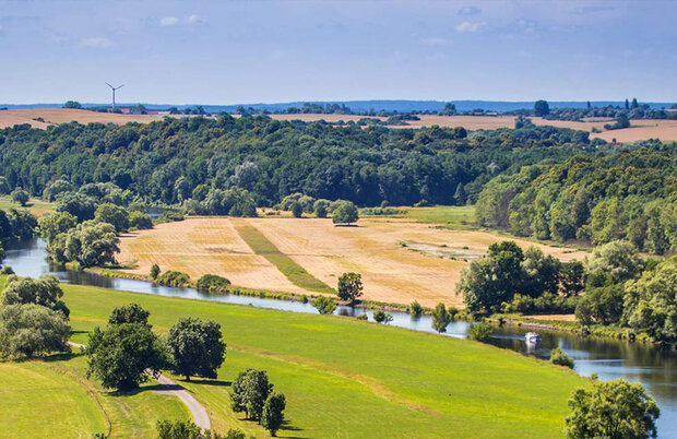 Fluss fließt durch grüne Wiesen und Felder mit Wald im Hintergrund unter blauem Himmel.