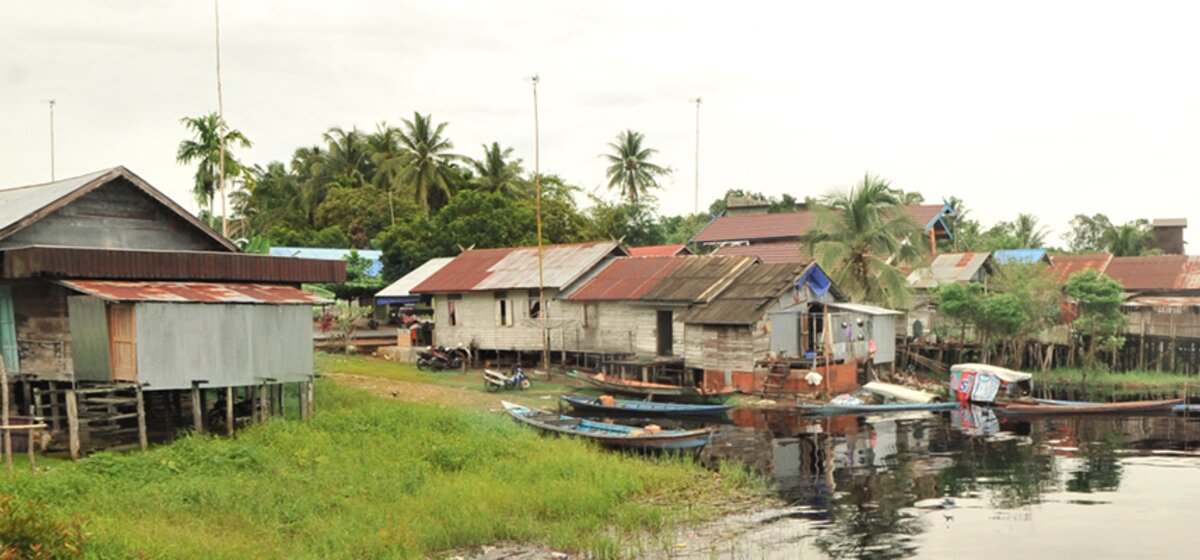 Hütten aus Holz und Wellblech stehen an einem Flussufer. Im Wasser liegen Boote.