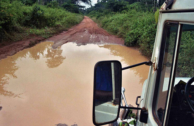 Unbefestigte rote Erdstraße mit großer Wasserpfütze, gesehen aus dem Fenster eines weißen Geländewagens.