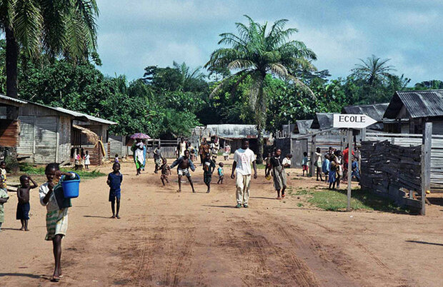 Kinder und Erwachsene gehen auf einem staubigen Weg in Dzanga Sangha mit einfachen Holzhäusern. im Hintergrund sind Bäume und Palmen.