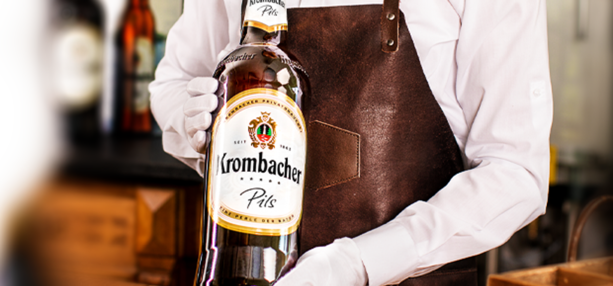 Person in weißem Hemd und brauner Schürze hält eine Krombacher Pils 3,0l in den Händen.