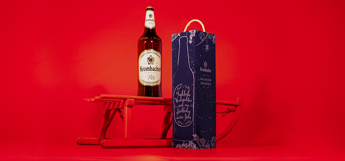 Eine Flasche 'Krombacher Pils' steht auf einem roten Schlitten. Daneben befindet sich eine Geschenktüte mit winterlichem Design und dem Text 'Frohe Weihnachten und ein gutes neues Jahr'.