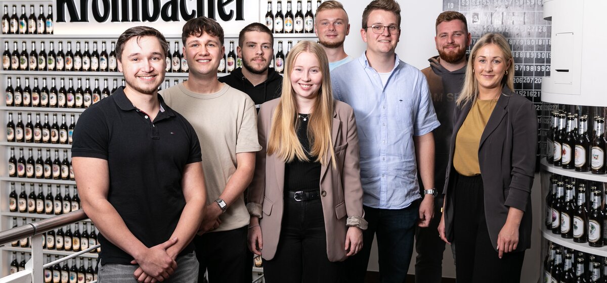 Acht lächelnde junge Männer und Frauen stehen vor einer Wand mit Krombacher Bierflaschen und einem großen Krombacher Schriftzug.