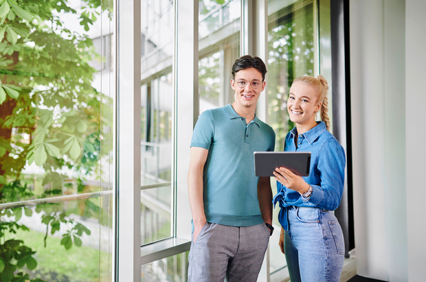 Ein junger Mann mit dunkelbraunen Haaren und einer Brille und eine junge blonde Frau, stehen nebeneinander und lächeln. Sie hält ein Tablet in der Hand.