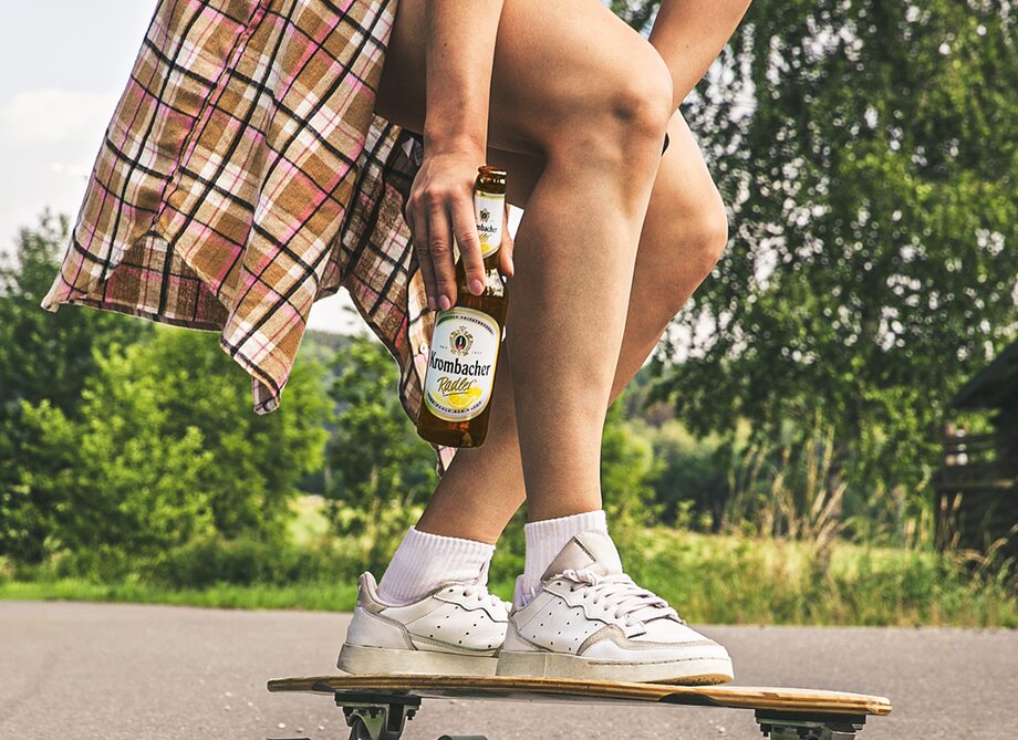 Auf einem Skateboard steht eine Frau, die sich hinhockt und eine Krombacher Radlerflasche in der Hand hält.