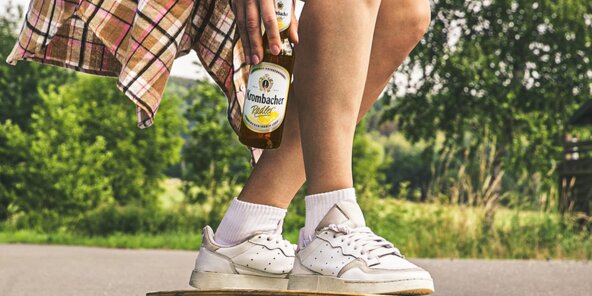 Auf einem Skateboard steht eine Frau, die sich hinhockt und eine Krombacher Radlerflasche in der Hand hält.