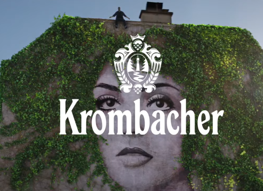 Ein Mann steht auf dem Dach eines Gebäudes, das mit grünen Pflanzen bewachsen ist. Im Vordergrund ist das Logo 'Krombacher' deutlich sichtbar.