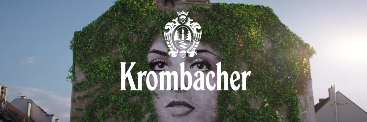 Ein Mann steht auf dem Dach eines Gebäudes, das mit grünen Pflanzen bewachsen ist. Im Vordergrund ist das Logo 'Krombacher' deutlich sichtbar.