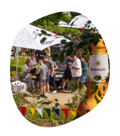 Menschen stehen unter Sonnenschirmen mit Krombacher-Logo beim Krombacher Sommerfest im Garten, bunte Wimpel und Blätter hängen im Vordergrund
