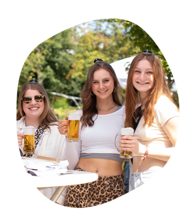 Drei junge Frauen stehen im Freien, halten Krombacher Seidel Biergläser und tragen ganz kleine Krombacher Schützenfest Hüte, eine trägt ein weißes Crop-Top und Leopardenmuster-Hose.