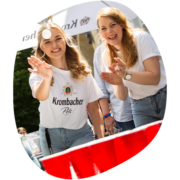 Zwei junge Frauen spielen Beer-Pong, eine wirft einen Ball, die andere klatscht, beide tragen weiße Krombacher T-Shirts und Jeans