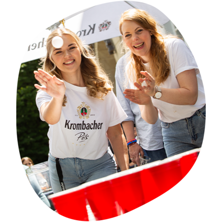 Zwei junge Frauen spielen Beer-Pong, eine wirft einen Ball, die andere klatscht, beide tragen weiße Krombacher T-Shirts und Jeans