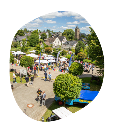 Sommerfest mit vielen Menschen, weißen Sonnenschirmen und bunten Wimpeln vor der Krombacher Erlebniswelt, im Hintergrund das Dorf mit Kirche und Wohnhäusern