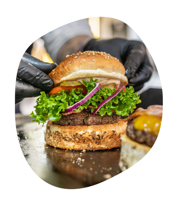 Frisch zubereiteter Hamburger mit Salat, Tomate und roten Zwiebeln, gehalten von Händen mit schwarzen Handschuhen