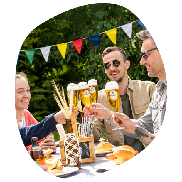 Vier Personen stoßen mit Krombacher Biergläsern an, im Hintergrund bunte Wimpel, auf dem Tisch Bratwurst im Brötchen, eine Flasche Krombacher Spezi, Deko und ein Besteckkorb.