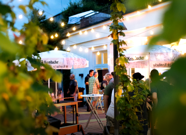 Abendliche Szene mit Menschen unter Lichterketten und Krombacher-Sonnenschirmen im Außenbereich der Krombacher Erlebniswelt.