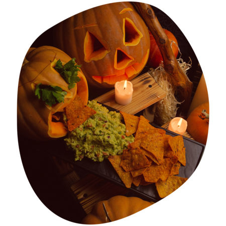 Zwei ausgehöhlte Kürbisse, einer mit geschnitztem Gesicht, der andere mit Guacamole und Tortilla-Chips gefüllt, umgeben von Kerzen und weiteren Kürbissen.