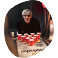 Person mit grauen Haaren lehnt sich über einen Tisch mit zehn roten Plastikbechern, die in einem Dreieck angeordnet sind. (Bierpongtisch)