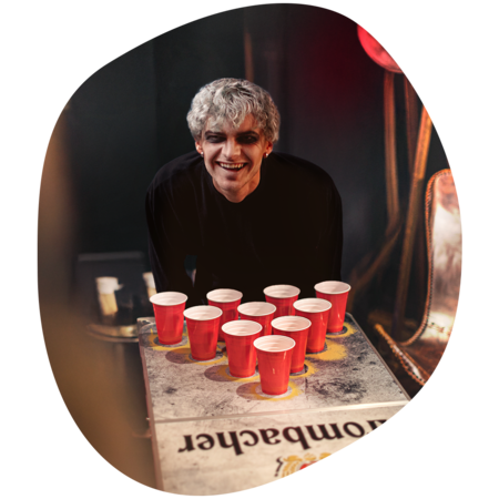 Person mit grauen Haaren lehnt sich über einen Tisch mit zehn roten Plastikbechern, die in einem Dreieck angeordnet sind. (Bierpongtisch)