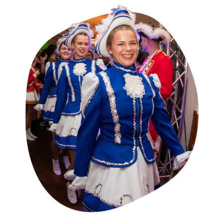 Frauen in blau-weißen Tanzkostümen mit weißen Stiefeln und Federhüten in einer Reihe, im Hintergrund Personen in roten Uniformen.