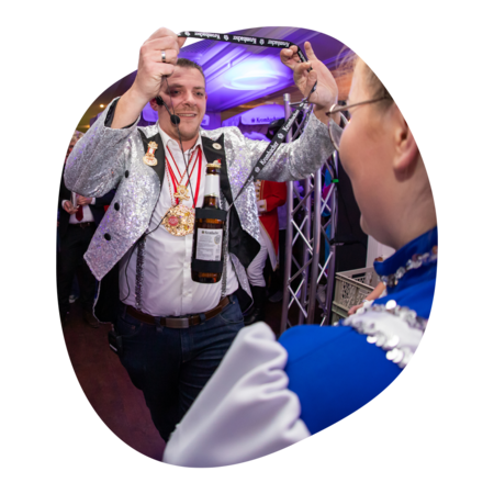 Person in silberner Jacke mit Medaillen hält eine Flasche mit schwarzem Etikett an einem Band hoch, im Vordergrund Person in blau-weißer Tracht.