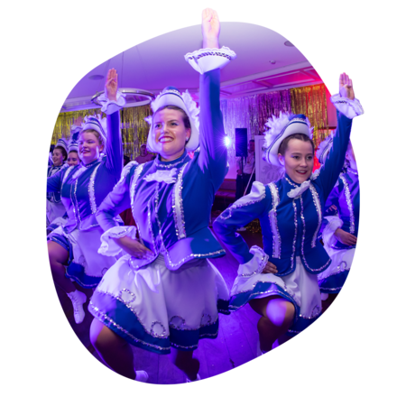 Tanzgruppe in blau-weißen Uniformen mit Rüschen und Mützen hebt synchron einen Arm und ein Bein bei Bühnenauftritt.