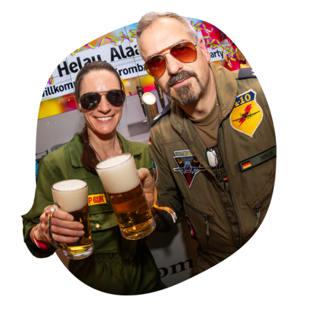 Zwei Personen in grüner und brauner Uniform mit Aufnähern halten Biergläser mit Schaum, im Hintergrund bunte Partydeko und Schriftzug 'Helau Alaaf'.