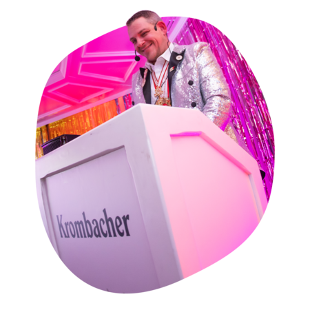 Person in silberner Glitzerjacke steht hinter einem weißen Pult mit der Aufschrift 'Krombacher', im Hintergrund pinke und orangefarbene Dekoration.