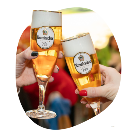 Zwei Hände mit unterschiedlich lackierten Fingernägeln halten Krombacher Pils in schlanken Biergläsern mit Schaumkrone.