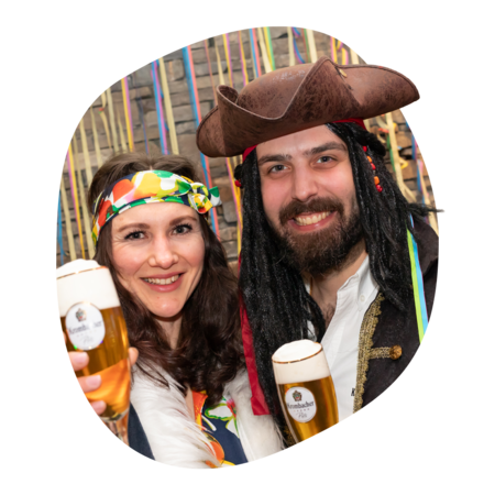 Zwei Personen in bunter Kleidung, eine mit Piratenhut und Dreadlocks, halten Gläser mit Krombacher Pils im Glas vor buntem Hintergrund.