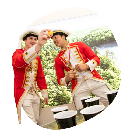 Zwei Männer in roten Uniformjacken mit goldenen Knöpfen (Gardemänner) und weißen Hosen spielen mit einem gelben Ball vor einem Tisch mit schwarzen Bechern (spielen Beerpong).