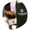 Person trägt grüne Krombacher-Schürze und hält ein Bratwürstchen in einem Brötchen mit schwarzen Handschuhen