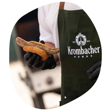 Person trägt grüne Krombacher-Schürze und hält ein Bratwürstchen in einem Brötchen mit schwarzen Handschuhen